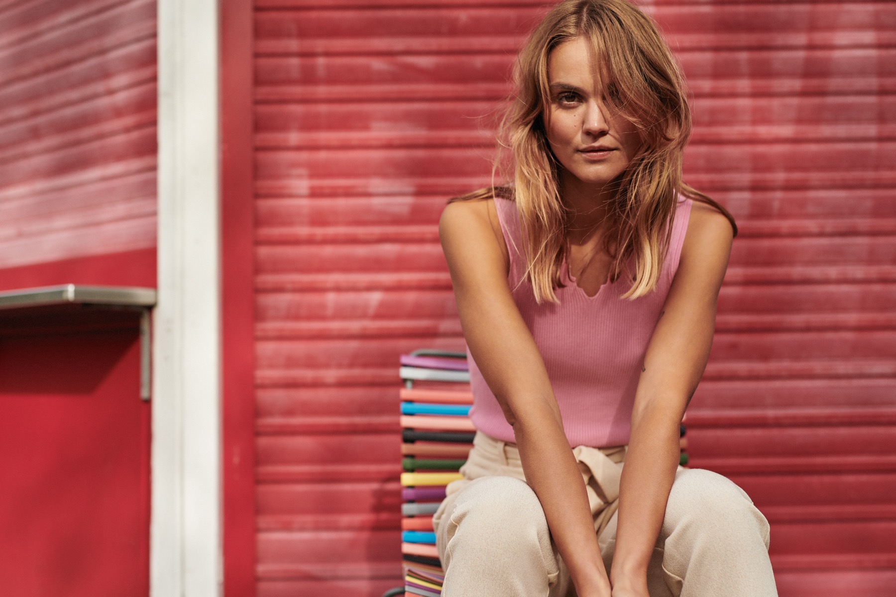 Young blonde woman in pink top sits on a colorful bench against a red wall.