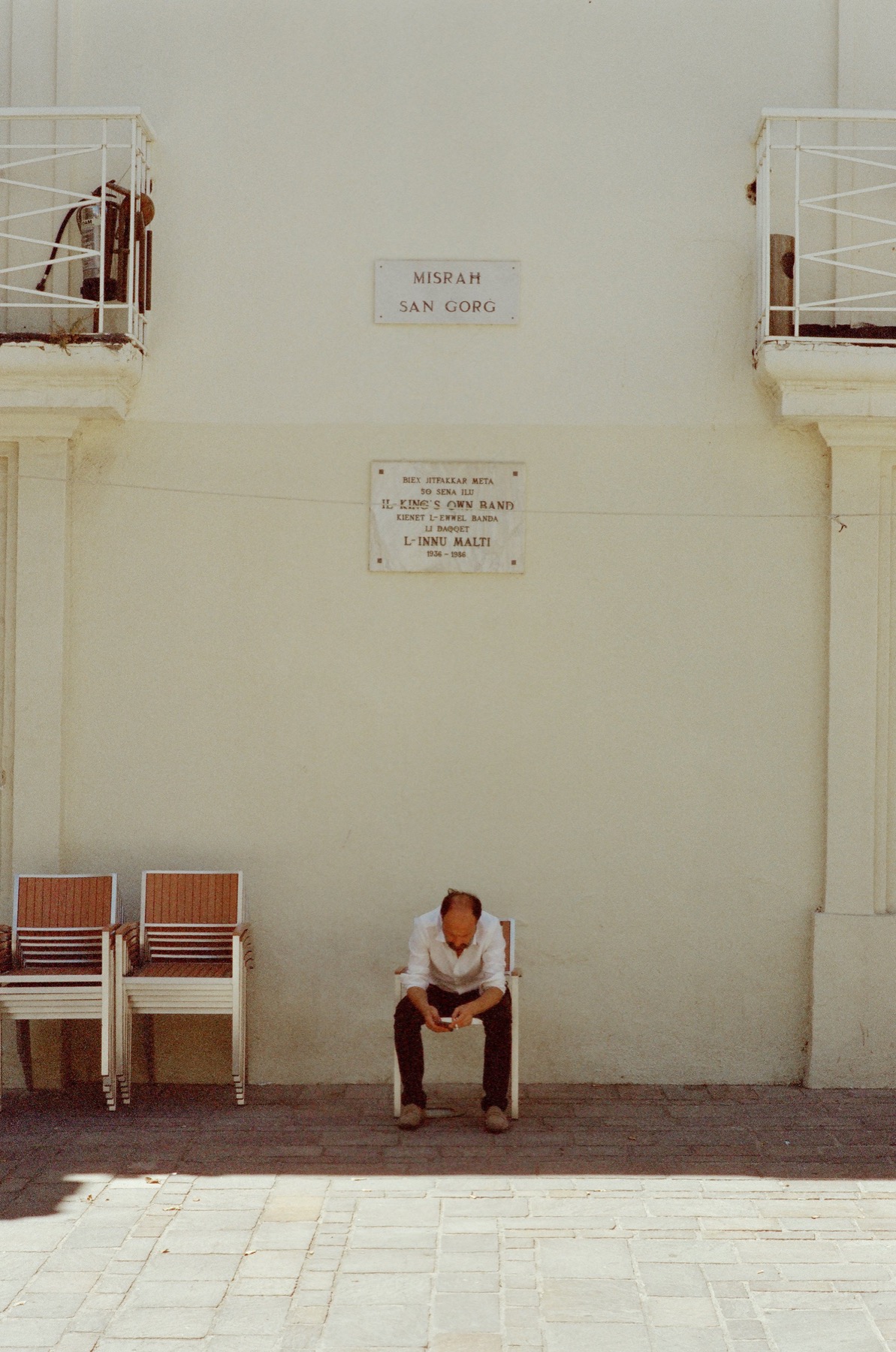 Man in a white shirt sits on a chair below wall plaques and balconies.