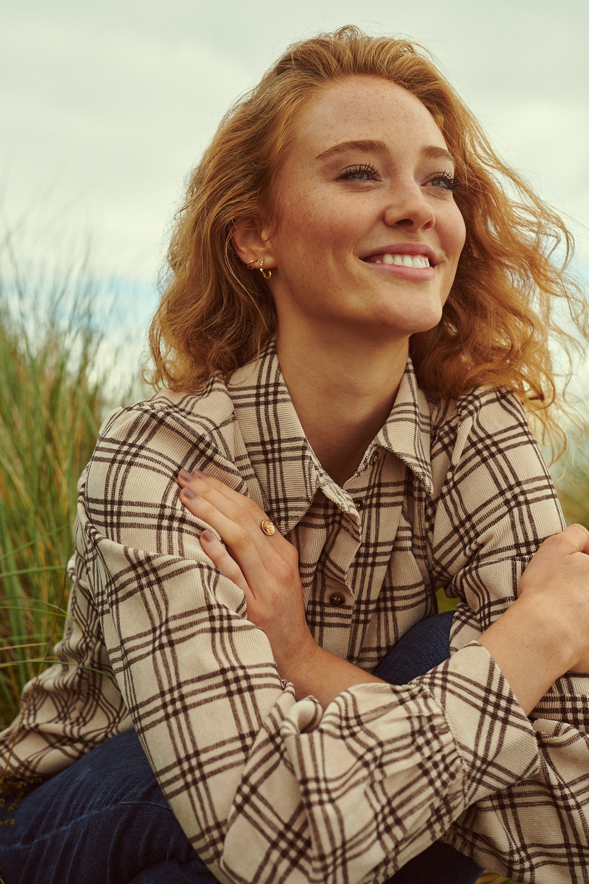 Red-haired woman with freckles smiling in a plaid shirt outdoors amidst tall grass.